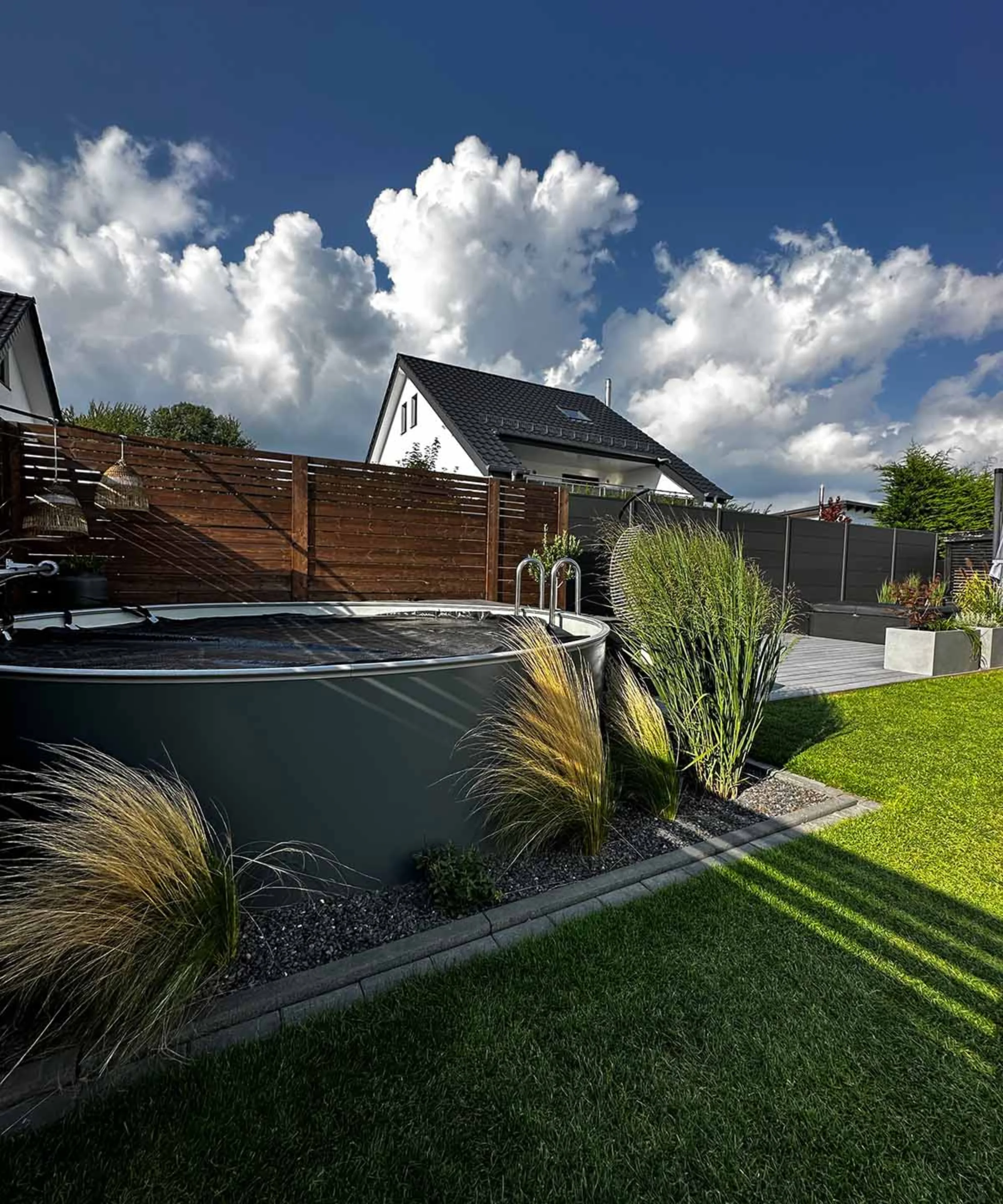 Ein moderner Garten mit Rasen, Ziergräsern und einem kleinen Pool unter blauem Himmel.