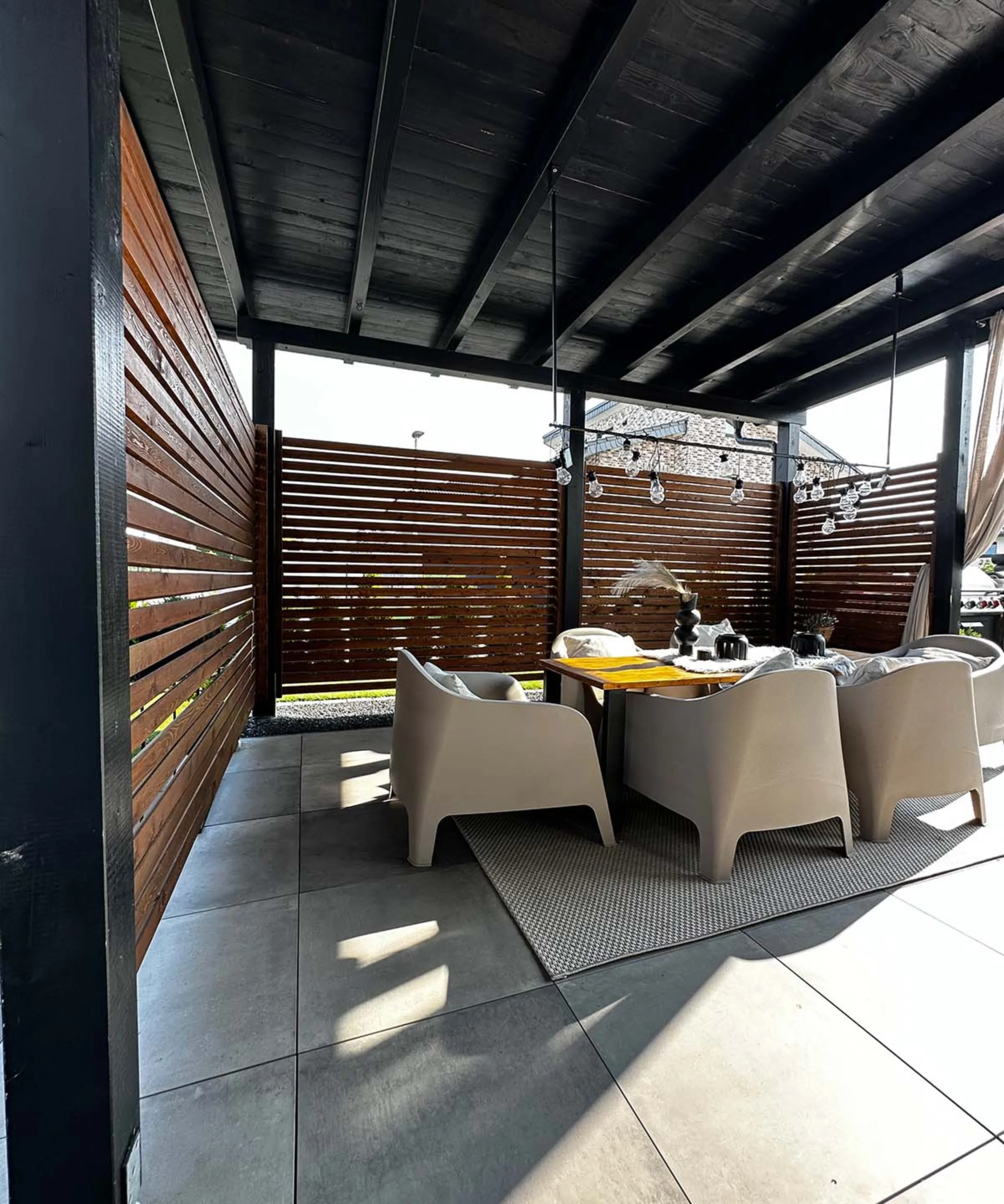 Eine überdachte Terrasse mit Tisch und vier Stühlen aus Rattan, umgeben von Holzlamellenwänden.